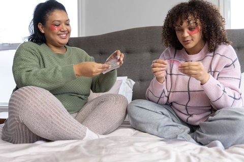 African american friends sharing pampering night with pink under-eye gel patches on bed
