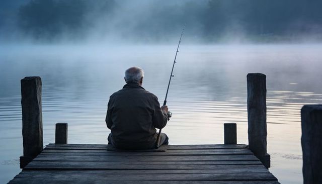 Senior man sitting on wooden dock fishing at misty dawn lake, peaceful solitary moment