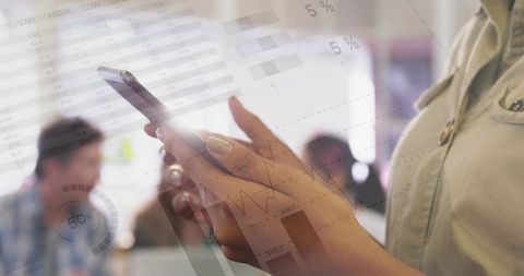 Hands tapping smartphone showing data and financial charts overlay in coworking workspace
