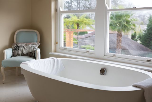 Modern bathroom featuring an empty white bathtub next to a large window with a view of greenery. A light blue upholstered chair with a decorative pillow is placed nearby. Ideal for use in articles or advertisements related to home decor, interior design, luxury living, or relaxation.