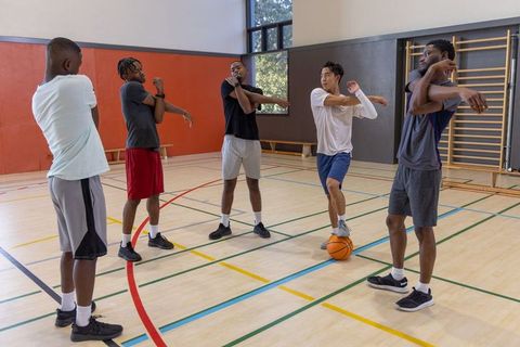 Diverse male athletes stretching together on indoor basketball court during warmup