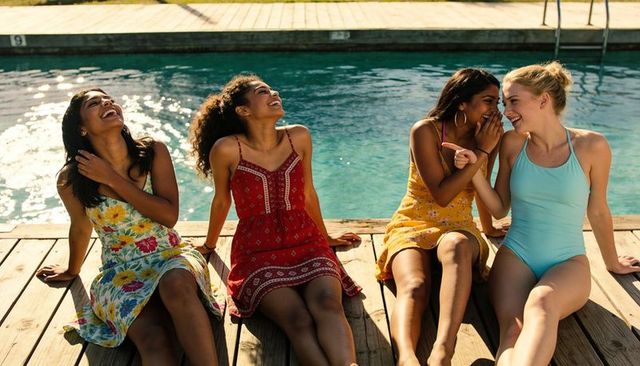 Four friends laughing and chatting poolside on deck wearing summer dresses and swimsuit
