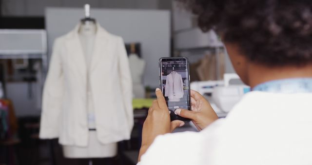 Fashion designer is taking a photograph of a white blazer using a smartphone in a well-organized sewing room. The focus is on capturing the blazer, which is situated on a mannequin. This image is perfect for depicting the everyday activities in the fashion industry, showcasing the creative process, and emphasizing the importance of technology in design. Ideal for articles, websites, or advertisements related to fashion designing, workshops, tailoring, or fashion technology.