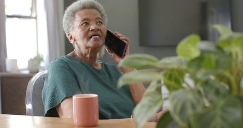 Senior african woman sitting at table talking on smartphone with mug and houseplant