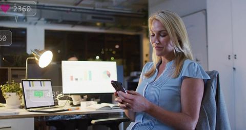 Blonde Woman Checking Phone While Reviewing Analytics at Night in Modern Open-Plan Office