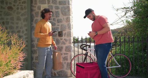 Courier and resident exchanging delivery while checking smartphones by bicycle on stone porch
