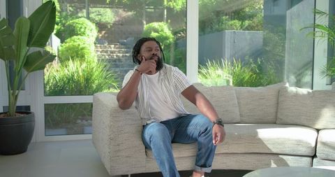 African man relaxing on modern light sofa in sunlit living room, looking thoughtful