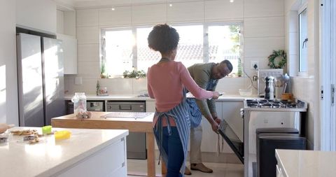 Couple baking in modern sunlit kitchen, man opening oven while woman arranging cookies