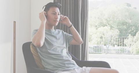 Asian man adjusting over-ear headphones relaxing in modern lounge chair near balcony