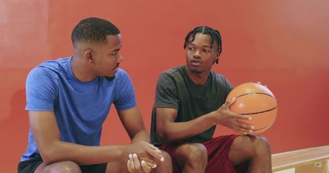Two african american men passing basketball and bantering on gym bench, showing teamwork