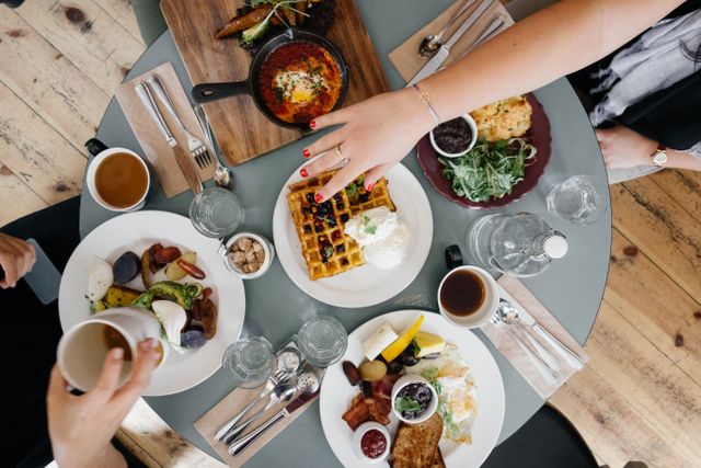 This image shows friends enjoying a brunch together with a variety of delicious dishes on a round table. The food includes waffles, salads, drinks, and other meals. Ideal for use in restaurant promotions, food blogs, social media posts, or advertisements centered around dining and social gatherings.