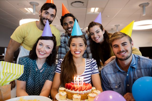 Diverse group of colleagues celebrating a birthday in an office environment. They are gathered around a table with a birthday cake adorned with candles and strawberries, wearing colorful party hats and smiling. Balloons and cupcakes add to the festive atmosphere. Ideal for use in articles or advertisements about workplace culture, team building, office celebrations, and employee engagement.