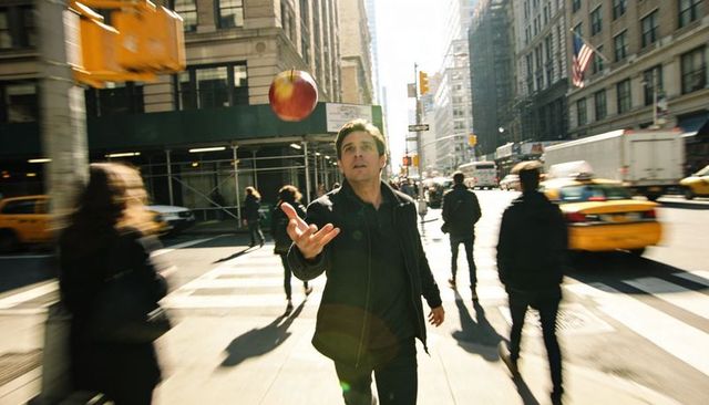 Man tossing apple on busy city crosswalk with motion blur, yellow taxis and sun flare