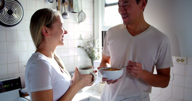 Couple is casually enjoying breakfast in a bright white kitchen. They are talking and smiling, with one person holding a coffee mug and the other a bowl of cereal. This could be used for concepts about morning routines, happy relationships, domestic life, or healthy lifestyles.