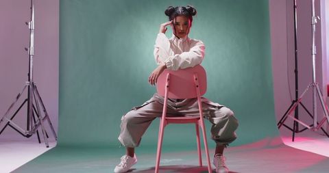 Edgy editorial studio portrait woman posing on pink chair wearing cargo pants and space buns