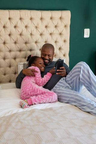 African-american father and daughter cuddling and laughing in bed in pajamas using phone