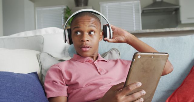 Teen boy sits on a comfortable couch, holding a tablet and wearing headphones in a contemporary living room. Ideal for illustrations of modern technology use, digital entertainment, and teenage lifestyle. Can be used in digital addiction awareness materials, tech product advertisements, and family living articles.