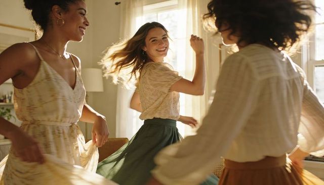 Young women twirling in sunlit living room wearing flowing skirts, smiling and laughing