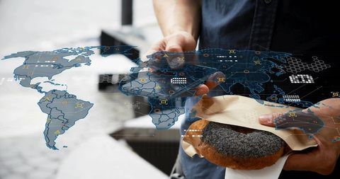 Asian man holding poppy-seed pastry tapping smartphone with holographic world map data overlay