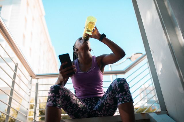 Fit african american woman resting on stairs, drinking water and using smartphone. healthy active lifestyle and outdoor fitness.