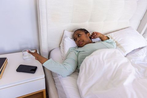 African american woman reaching for glass of water on bedside table while resting in bed