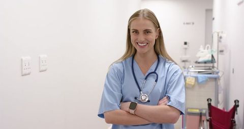 Smiling nurse in blue scrubs standing in hospital corridor with stethoscope and wheelchair