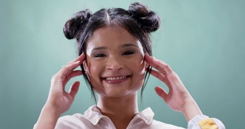 Smiling Asian woman raising hands to temples wearing rings and double buns studio portrait
