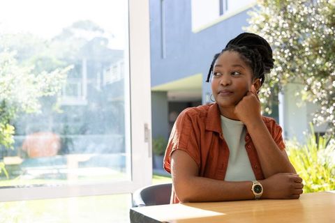 Contemplative african woman sitting at table gazing out to sunlit backyard pool relaxed home