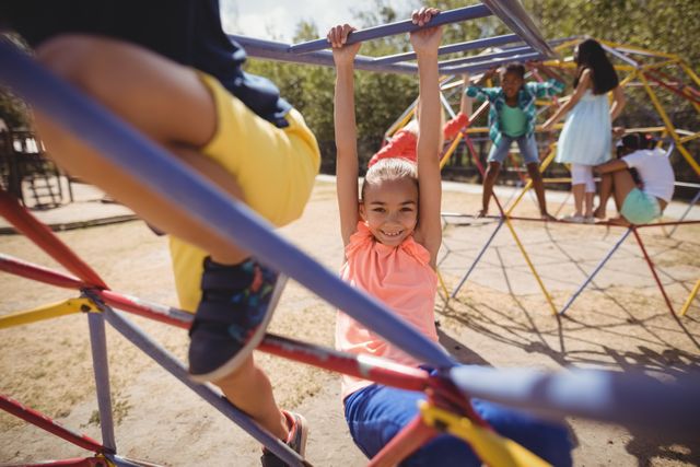 Happy girl hanging on monkey bars at a school playground with other children playing in the background. Ideal for use in educational materials, advertisements for playground equipment, or articles about childhood development and outdoor activities.