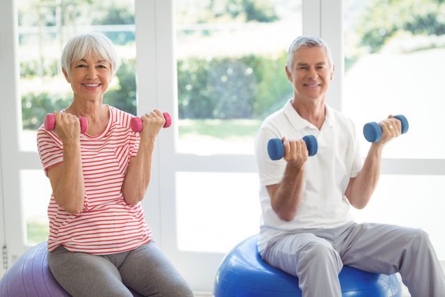 Senior couple lifting dumbbells while sitting on exercise balls in a bright home setting. Ideal for promoting senior fitness, home workouts, active lifestyle, and wellness among the elderly. Suitable for use in articles about fitness for seniors, healthy aging, and maintaining physical activity in later years.