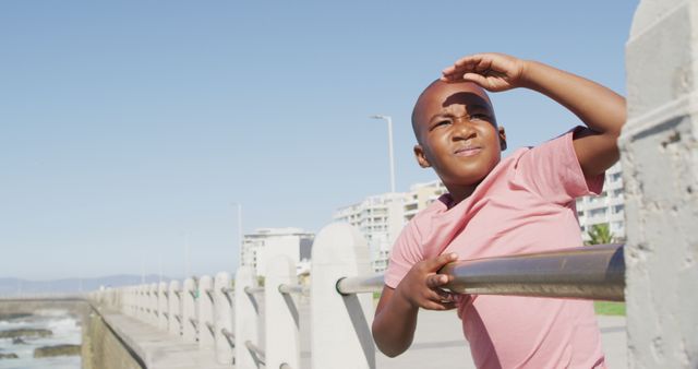 African American boy standing by a metal railing and ocean, shielding eyes while looking into the distance. Perfect for themes such as childhood, exploration, relaxing activities, and family vacations.