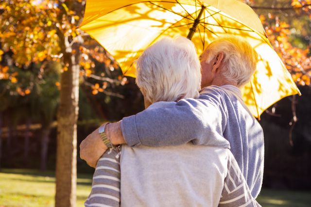Senior couple embracing under a yellow umbrella in a park during autumn. Ideal for themes related to love, companionship, retirement, and enjoying life in the golden years. Suitable for use in advertisements, brochures, and articles about senior living, health, and wellness.
