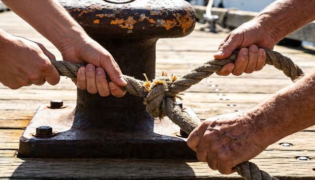 Hands tightening frayed mooring knot on sunlit wooden pier with rusted bollard, teamwork