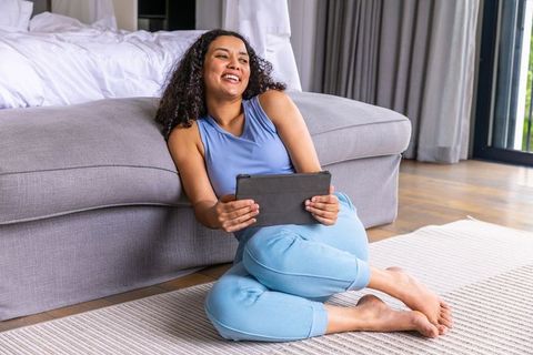 African american woman relaxing on bedroom floor holding tablet, cozy home leisure