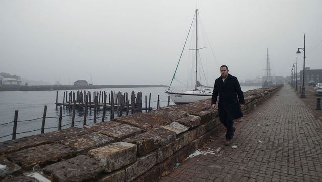 Man walking along foggy harbor seawall past moored sailboat on cobblestone promenade