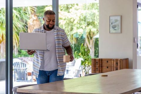 African american man holding laptop and mug in sunlit kitchen near patio doors, working remotely