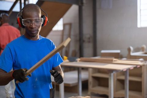 African american carpenter inspecting wooden plank wearing goggles and earmuffs in workshop