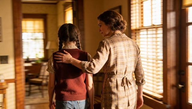 Mother guiding daughter down sunlit hallway, hand on shoulder, warm cozy vintage intimacy