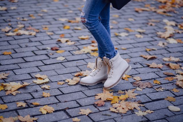 Featuring a closeup of a woman wearing distressed jeans and beige boots walking on a path covered with autumn leaves. Perfect for autumn fashion, lifestyle, outdoor activities, or nature-themed promotions.