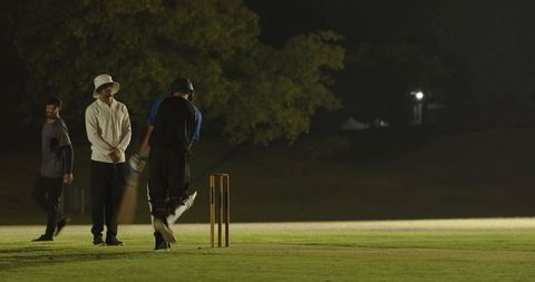 Night cricket practice under floodlights with batsman swinging and bowler returning