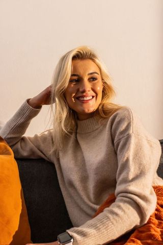 Smiling woman sitting on sofa wearing beige sweater and smartwatch in sunlit cozy home