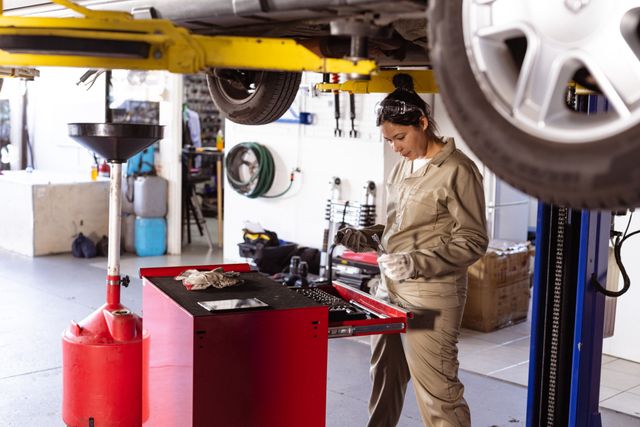 Asian female mechanic using tools in a car repair workshop. Ideal for illustrating automotive repair services, professional mechanics, and women in engineering. Useful for websites, brochures, and advertisements related to car maintenance, repair shops, and technical training programs.