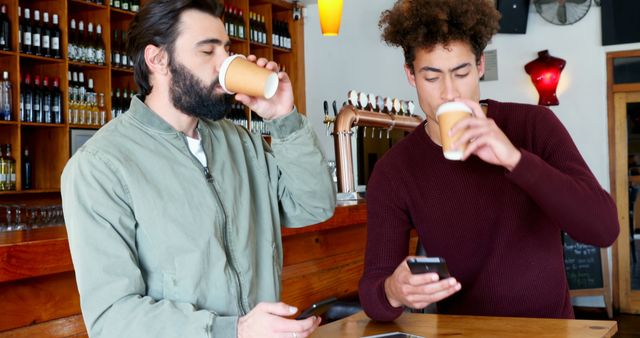 Two men are standing in a cafe, enjoying coffee from paper cups while using their smartphones. One man has a beard and is wearing a light green jacket, and the other has curly hair and is wearing a dark red sweater. This scenario illustrates modern social interaction and relaxation in a casual environment. Great for themes related to technology, coffee culture, digital communication, and social interaction.