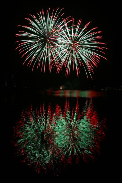 Spectacular red and green fireworks exploding in the night sky, with vibrant reflections on calm water. Perfect for illustrating festive celebrations such as New Year's Eve, Fourth of July, or other significant events. Ideal for festive promotions, holiday themes, event advertising, and celebratory contexts.