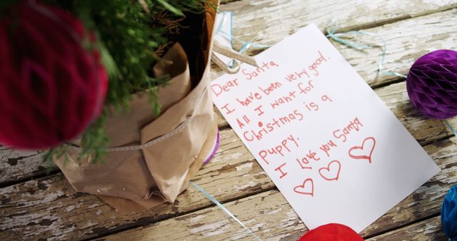 Handwritten Christmas wish list to Santa on wooden table next to potted plant and colorful holiday decorations. Ideal for holiday marketing, Christmas blog posts, nostalgic holiday cards, family tradition promotions, and festive imagery in holiday advertisements.