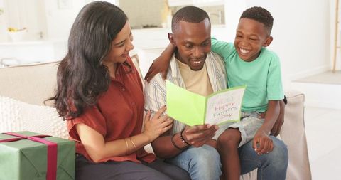 Father receiving happy father's day card from smiling son and partner on sunlit sofa