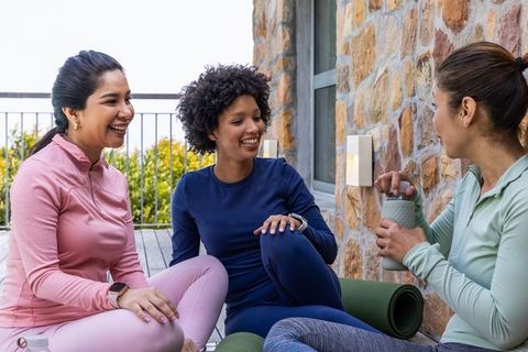 Three women in activewear sharing yoga mats and chatting on sunlit terrace after workout