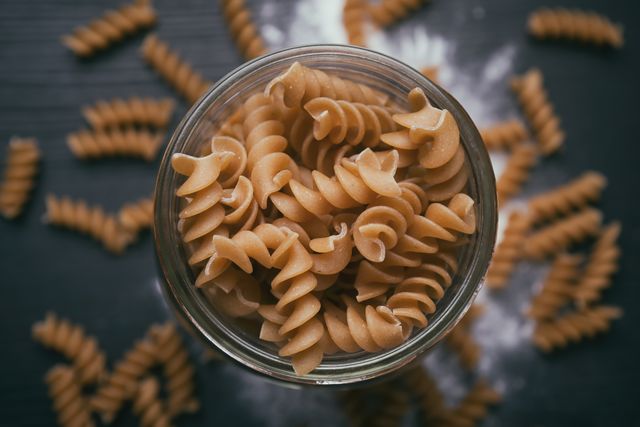 Top view of whole wheat fusilli pasta in glass jar on dark background scattered with pasta pieces. Perfect for food blogs, healthy eating promotions, or cooking websites.