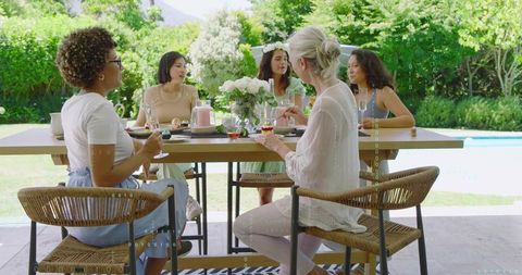 Women chatting around wooden table by pool at alfresco brunch, celebrating friendship