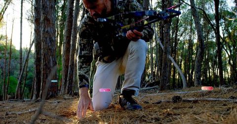 Kneeling hunter scanning pine forest floor with compound bow at golden hour tracking signs